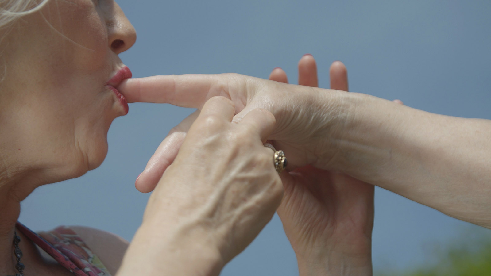 The picture shows the lower part of an older white woman's face and a white hand, whose index finger she is sucking. Her lips are painted pink.