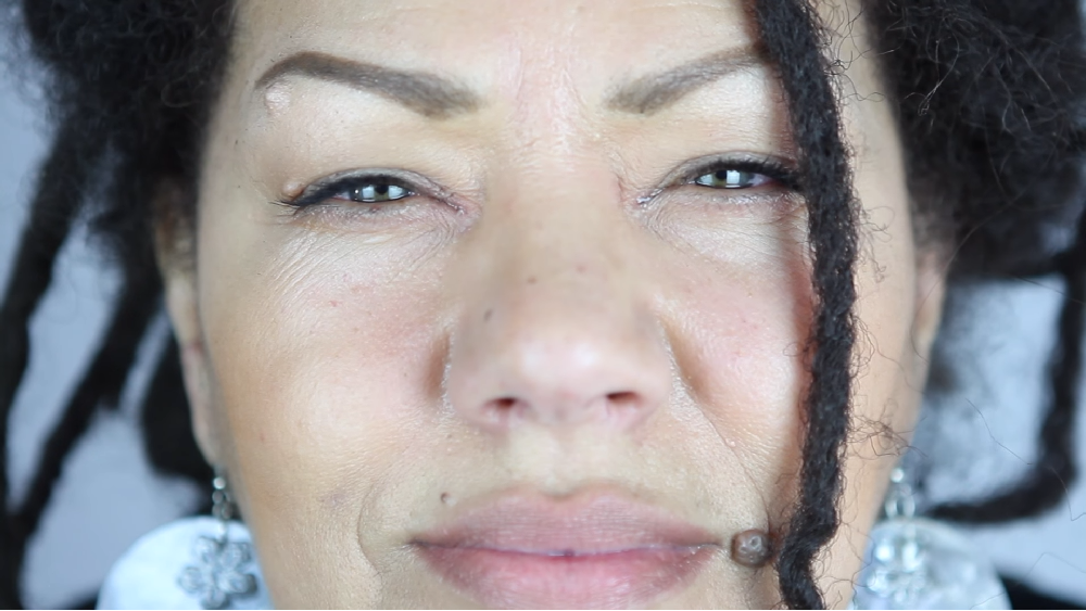 The face of a woman of colour looking directly in the camera is shown. She has dreadlocks, wears blue colourful earrings and tiny wrinkles can be seen.