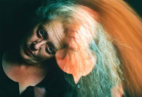 A white woman with long, grey hair looks at the camera with blue eyes and her head tilted. Double exposure shows her with her head up right out of focus.