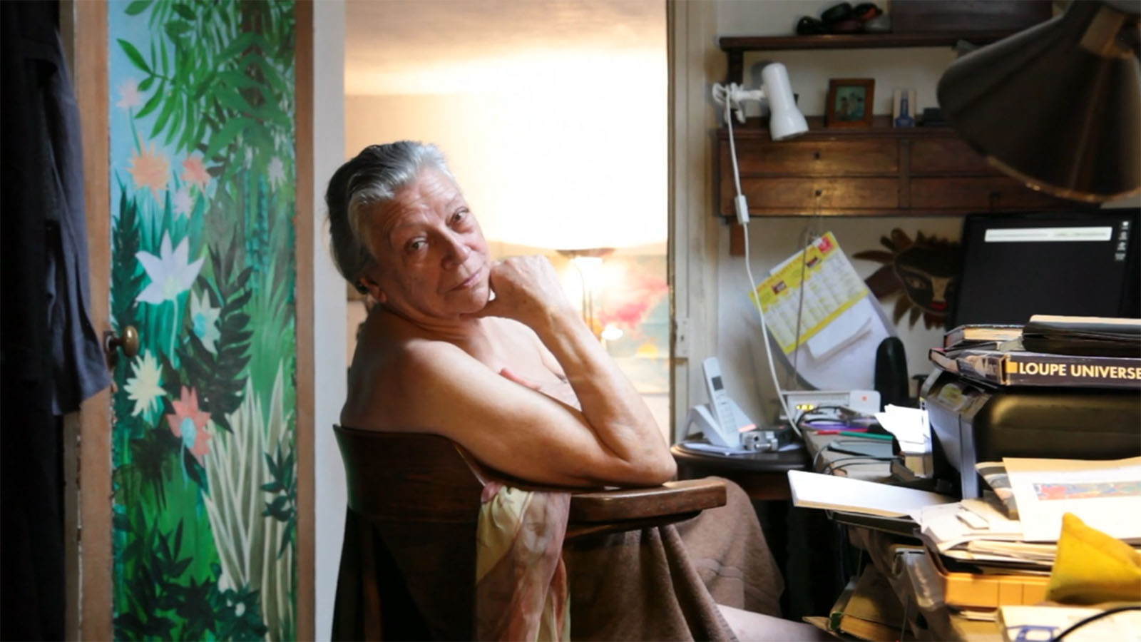 Thérèse is sitting in a wooden chair in an office. She is wearing a towel and looks back to the camera over her bare shoulder.