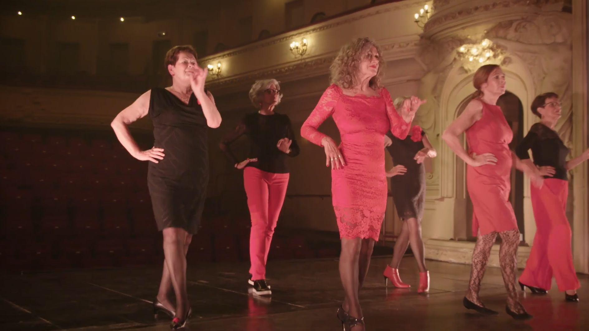 Six women dancing on a warmly lit stage. They wear feminine red and black clothing and are moving delicately