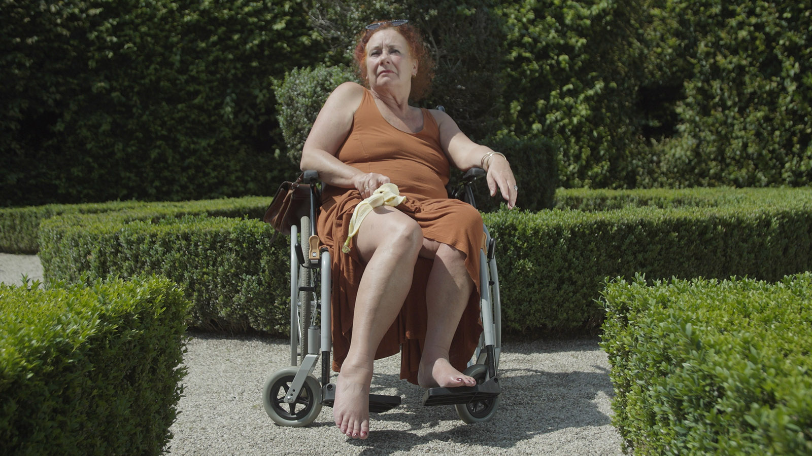Barefooted person in rust colored dress sitting in a wheelchair placed in a garden. One arm is holding a brown handbag and a half-peeled banana.