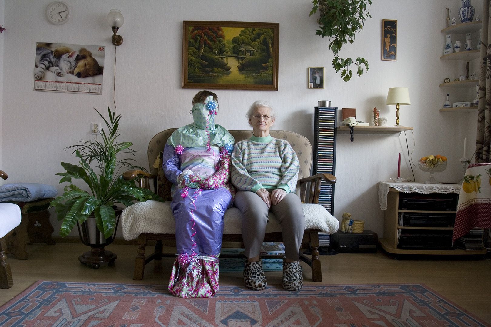 Two persons sitting on a sofa in a living room. The person on the left is covered in purple, pink, and blue wrapping paper and is adorned with bows.