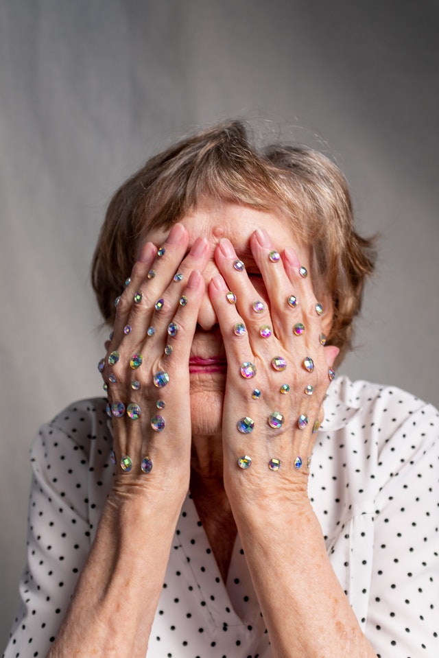 An aged person in a black-and-white polka dot blouse hiding their face with their hands covered in rhinestones.