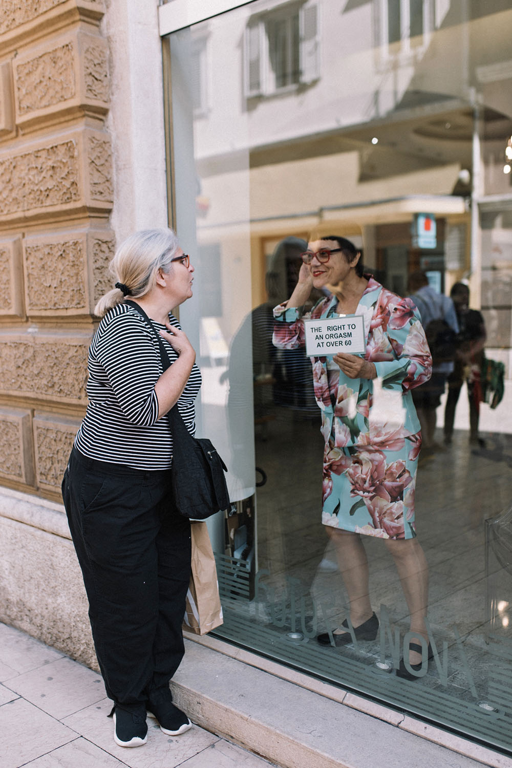 The artist is standing in a shop window holding a sign with the project's title, interacting with an older woman on the street outside.